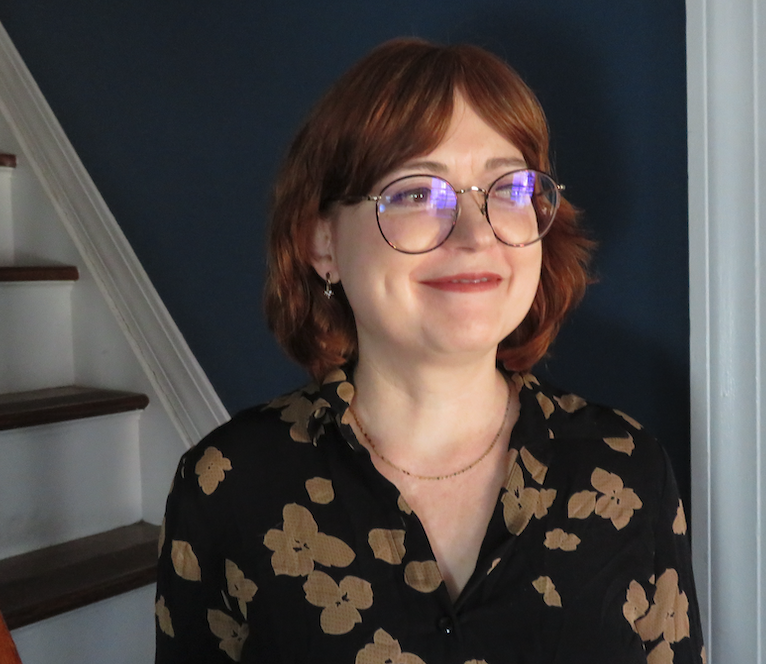 Maud Newton stands in front of stairs, smiling in a black shirt with a brown flower print on it